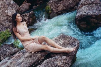 Young woman in the dress is sitting on the stone in the middle of a creek. Fashionable toning.