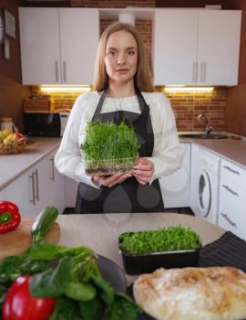 A young blonde happy lady standing in the kitchen while cooking healthy food. Healthy Lifestyle Concept. Cooking at home. microgreens in a container as a guarantee of healthy intestinal microflora