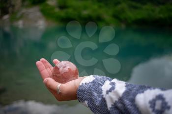 A woman's hand holds a spherical glass ball against the background of a beautiful mountain lake. Turier lake, Alibek glacier, North Caucasus, Dombai,, Russia.