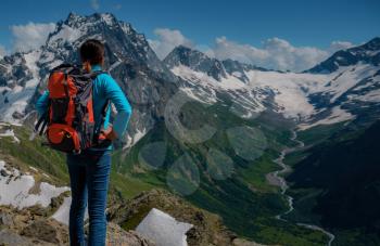 A young woman tourist in a blue blouse with an orange hiking backpack enjoys a breathtaking view of the mountains. North Caucasus, Dombai, Russia. mountaineering sport lifestyle concept