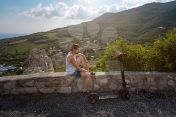 Young man riding an electric scooter on mountain range. Rest after a long trip, enjoying fresh air and beautiful scenic views. Ecological transportation concept