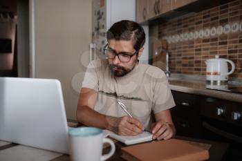 Concentrated millennial hipster guy wearing glasses, listening to favorite music while planning workday. Focused young biracial businessman watching educational lecture. writing down notes.