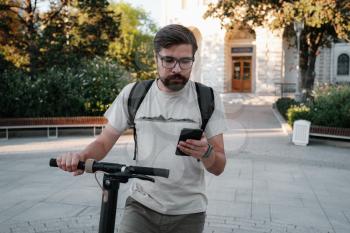 hipster man commuter with electric scooter outdoors in city, using smartphone. Young millennial guy using cellphone rental of eco transportation in summer
