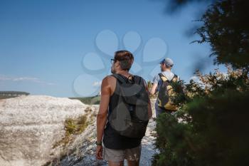 Two Men walking on rocky slope carrying Backpacks using trekking Sticks. Mountains valley View beside of People. summer travel to nature. Hiker in background.