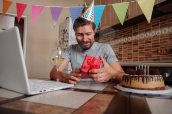 Man celebrating birthday online in quarantine time. Coronavirus outbreak 2020. The guy opens the box and is very happy with the gift. Communicating with friends remotely