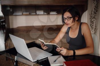 Young asian female student in glasses using laptop, read a book. Cozy office workplace, remote work, E learning concept.