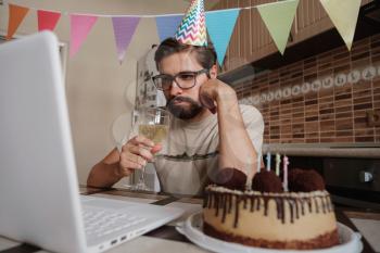 Upset young man sitting at the birthday cake and looking with sad eyes on it. Concept of solitude in quarantine during the Coronavirus Pandemic COVID-19.