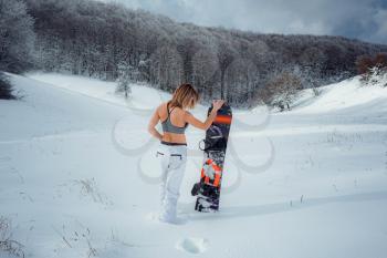 Female snowboarder hold snowboard and going to snowboarding. winter sport activity, forest snow outdoors lifestyle. Girl wearing a short top and ski white pants.