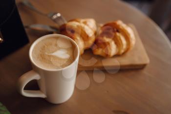 Cappuccino with beautiful latte art and croissant on wooden background on the table. Perfect breacfast in the morning. Rustic style.