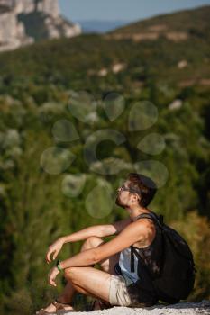 Young male hiker with backpack relaxing on top of a mountain during calm summer sunset - scenery from vacation. Tired traveling man resting on hill.