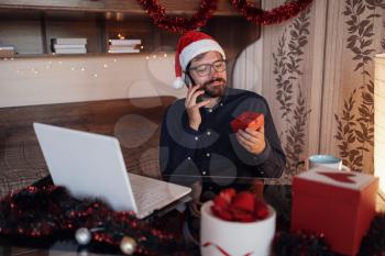 Close up portrait of joyful Caucasian handsome man in santa hat sitting in cozy room xmas decoration calling by smartphone with happy face . New Year. Merry Christmas