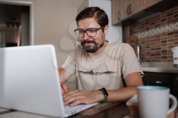 Focused young man wearing glasses using laptop, typing on keyboard, writing email or message, chatting, shopping, successful freelancer working online on computer, sitting in kitchen