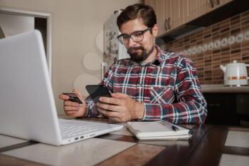 Handsome man using credit card to pay online with smartphone. Shopping Online, Banking system. guy in a plaid shirt sitting in his home kitchen