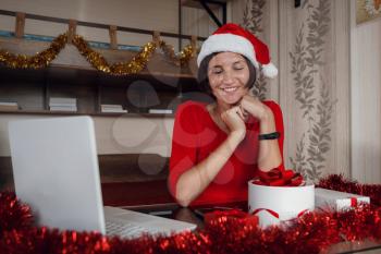 A happy woman in a red shirt is sitting in front of a computer screen with a gift box in her hands. Remote Christmas video party. Family quality time during pandemic. Online video call conferencing.