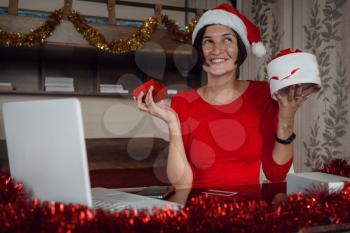 A happy woman in a red shirt is sitting in front of a computer screen with a gift box in her hands. Remote Christmas video party. Family quality time during pandemic. Online video call conferencing.