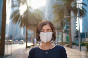 Portrait of young woman wearing in blue dress and white mask for prevent virus, walk in front of skycrapers in modern city.