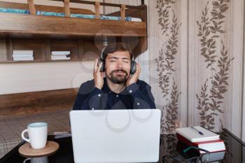 Satisfied businessman wearing headphones enjoying music with closed eyes, relaxing during break, sitting at work desk, smiling young man wearing glasses listening to favorite song