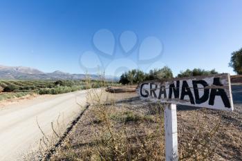 Road sign to Granada by side of dry dusty road with olive trees and mountains in the distance