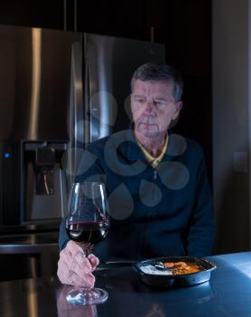 Lonely and depressed senior male sitting alone at kitchen table eating a microwaved ready meal of curry with red wine