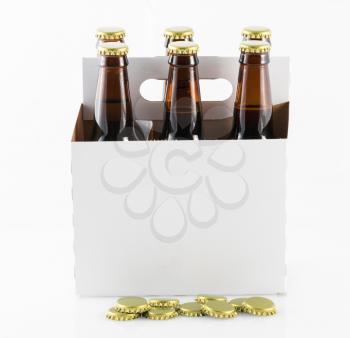 Six beer bottles in cardboard container with gold caps with side of carrier facing camera and bottle caps on table