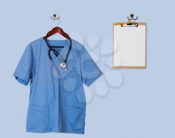 Blue medical scrubs uniform shirt hanging on a hanger with stethoscope and on hanger and hook on blue wall with clipboard for message