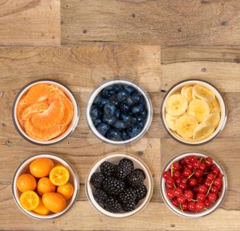 Top view of a white glass bowls of various sorts of organic fruits including, banana, orange, blueberry, redcurrant, blackberry and kiwi berry and sitting on old wood table surface