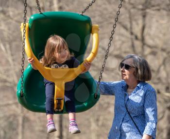 Grandma with her young granddaughter playing in a modern plastic swing in forest playground