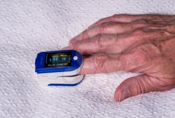 Pulse oximeter on finger used to test blood oxygen level in case of virus infection of the lungs with senior hand resting on bedspread