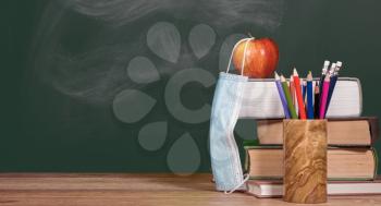 Concept for back to school with coronavirus or Covid-19 with books and apple against a chalkboard with face mask