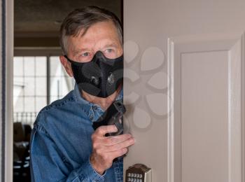 Senior man with face mask looking worried at front door and ready to defend home with a gun during coronavirus quarantine