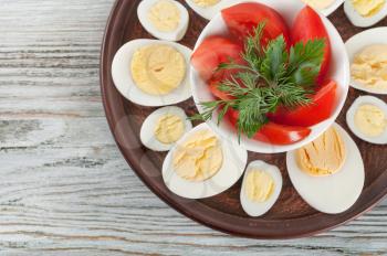 Boiled hen eggs and red tomato in a clay dish on a wooden background. Top view.