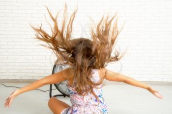 The little girl is sitting in front of the fan and is cooling. Air streams her hair