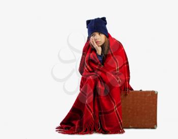 A little girl is waiting for a trip to the warm lands, sitting on a suitcase with luggage, but while she is wrapped in a woolen blanket from the cold