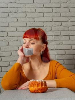 Conceptual image of a young red-haired girl on a diet sitting with her mouth taped with silver tape before appetizing pastries