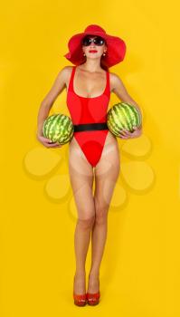 young girl in a bright red swimsuit and a wide-brimmed red hat eats a watermelon on a yellow background