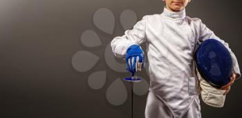 little boy fencer armed with a rapier in white sports equipment and with a protective helmet on a dark background