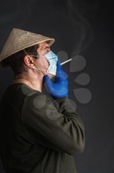 Chinese adult man in traditional Asian conical straw hat smokes a cigarette in a virus-protective medical mask on a dark background