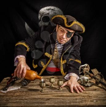 An adult pirate captain drinks alcohol from a clay bottle in a glass against the background of a black flag Jolly Roger