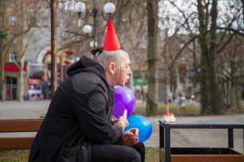 A sad birthday boy wearing a homemade party cap with balloons and cake with a candle celebrates his birthday alone in an empty park during an epidemic