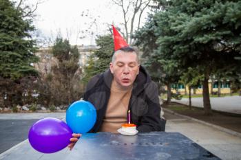 A sad birthday boy wearing a homemade party cap with balloons and cake with a candle celebrates his birthday alone in an empty park during an epidemic