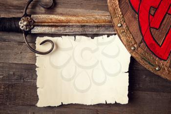 Vintage medieval background with sword and shield and blank sheet of blank paper or card on rough wooden table