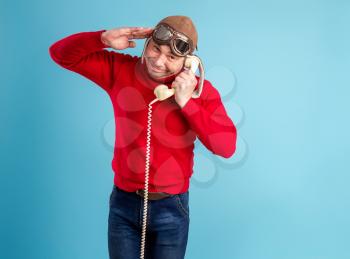 An adult man in a pilot's helmet and glasses communicates playfully and cheerfully on an old phone