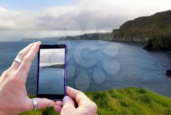 View over the mobile phone display during taking a picture of coast and ocean in Northern Ireland. Holding the mobile phone in hands and taking a photo. Focused on mobile phone screen. 