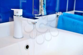 White sink faucet closeup with flowing water in bathroom with blue tiles.