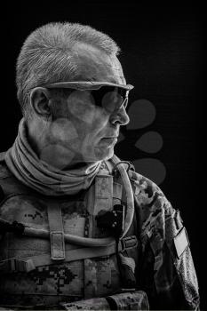 Black and white studio shot of special forces white-haired veteran in field uniforms, black background. Protective goggles glasses are on