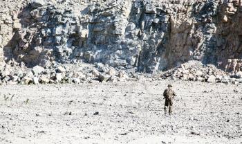 Location shot of soldier in field uniforms with rifle moving in the desert among rocks. Back view