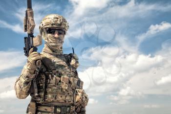 Half-length portrait of airsoft game player in army camouflage uniform, tactical helmet, load carrier and face hidden behind mask, posing with firearm replica in hands, cloudy sky on background