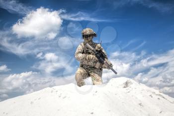 Man in military camouflage uniform with service rifle replica, standing on top of sand dune with cloudy sky on background, imitating U.S. army special forces shooter during airsoft war games in desert