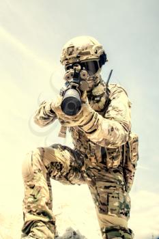 Airsoft war game player in army combat camouflage uniform, protected with helmet and tactical glasses, aiming in camera with optical sight on service assault rifle replica while climbing on sand dune