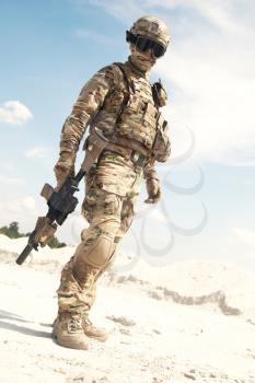 Airsoft player, military games participant in U.S. army infantry camouflage uniform, tactical mask, helmet and glasses, standing in desert area with combat service rifle or carbine replica in hands